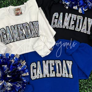 May include: Three sweatshirts in white, black, and blue with the word "GAMEDAY" in silver glitter letters. The sweatshirts are laid out on a green grassy surface with a silver and blue pom-pom.