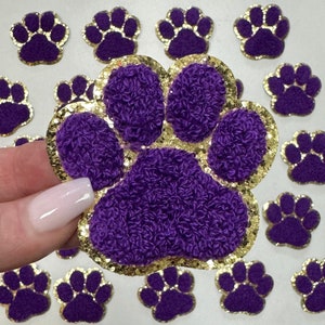 May include: Purple and gold glitter paw print iron-on patches. The paw prints are fuzzy and have a gold glitter outline.