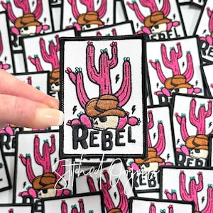 May include: A pink and green embroidered iron-on patch with a skull wearing a cowboy hat and the word "Rebel" in black lettering. The skull is surrounded by cactus plants.
