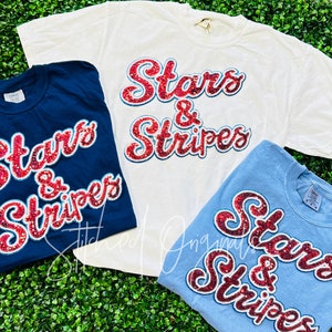May include: Three t-shirts with the phrase "Stars & Stripes" in red and blue sequins. The shirts are white, navy blue, and light blue.