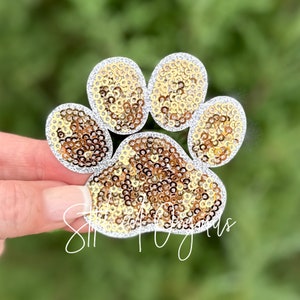 May include: A gold and silver sparkly paw print iron-on patch. The paw print is made of sequins and has a silver outline.