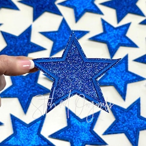 May include: A close-up of a blue glitter star-shaped patch. The patch has a double stitched outline and is surrounded by other blue glitter star-shaped patches.