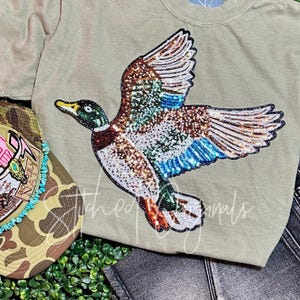 May include: A light green t-shirt with a sequined duck appliqué. The duck is flying with its wings spread and has a blue, green, and brown color scheme.