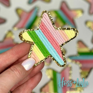 May include: A rainbow striped star-shaped patch with a gold glitter outline. The patch is made of fabric and has a stitched design. The text "Hoss + Lula Stitched" is visible in the bottom right corner of the image.