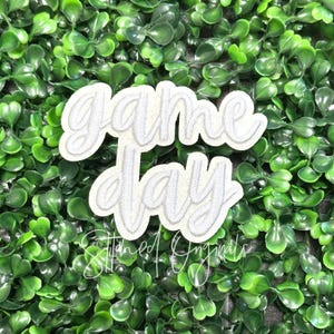 May include: A white embroidered patch with the words "Game Day" in a cursive font. The patch has a light gray outline and is set against a backdrop of green foliage. The text "Stitched Originals" is at the bottom.