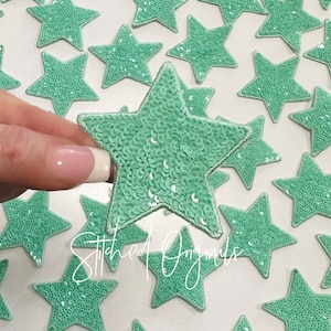May include: A collection of mint green star-shaped fabric patches with sequins. The patches are approximately 2 inches in diameter and have a stitched edge.