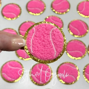 May include: Pink tennis ball with a white seam, surrounded by a gold glitter trim. The tennis ball is fuzzy and has a stitched design. The background is white.