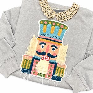 May include: A light gray sweatshirt featuring a large, colorful nutcracker patch with textured yarn details. The nutcracker has a blue hat, pink face, dark mustache, and green and pink uniform. A detachable white pearl collar is attached at the neckline.
