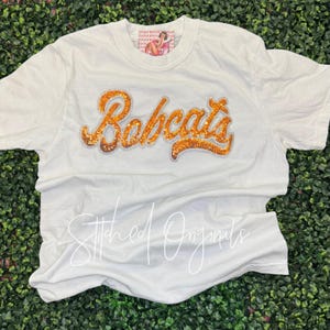 May include: A white short-sleeve t-shirt featuring the word Bobcats in orange sequined script lettering across the chest. The shirt includes a small rectangular brand label at the inner neckline with a graphic illustration of a person.