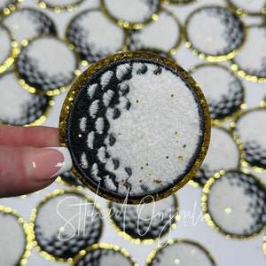May include: Embroidered golf ball patch with a gold glitter border. The golf ball design features a white textured surface and black and white stitched details. The patch is held by a hand, with other golf ball patches in the background.