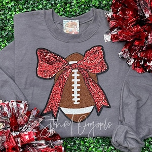 May include: A gray sweatshirt with a brown football and a red sequined bow. The football has white laces and the bow is tied around the football. The sweatshirt has the text "Stitched Originals" on the bottom.