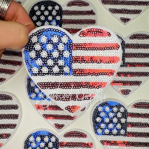 May include: Heart-shaped sequin patches in red, white, and blue, resembling the American flag. The patches feature a blue field with white stars and red and white stripes. The patches are on a white background.
