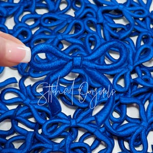 May include: A close-up of a pile of blue fabric bows. The bows are all the same size and shape, and they are arranged in a random pattern. The bows are made of a soft, velvety fabric.