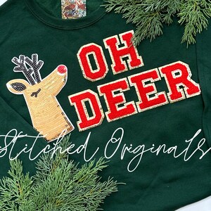 May include: A green sweatshirt with the text "OH DEER" in red and gold letters. A sparkly reindeer with a red nose is on the left side of the sweatshirt. The text "Stitched Originals" is at the bottom of the sweatshirt.
