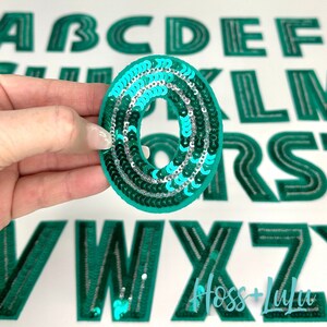May include: A teal green letter O made of sequins. The letter is surrounded by other teal green letters of the alphabet.
