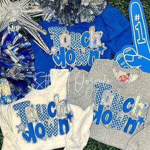 May include: A collection of sweatshirts in white, blue, and gray, featuring the text "Touch Down" in a checkered and star design. Includes a blue foam finger and pom-poms, perfect for a football fan.