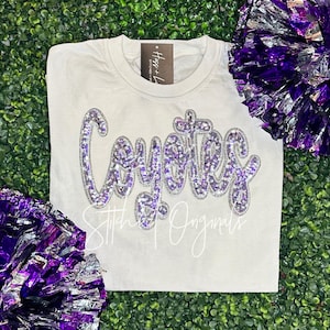 May include: A white t-shirt with the word "Coyotes" in large, sequined, purple letters. The shirt also features the text "Stitch Originals" in a cursive font. Purple and silver pom-poms are on either side of the shirt.