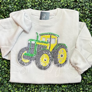 May include: A white sweatshirt with a green and yellow tractor design made of sequins. The tractor is a popular farm vehicle.