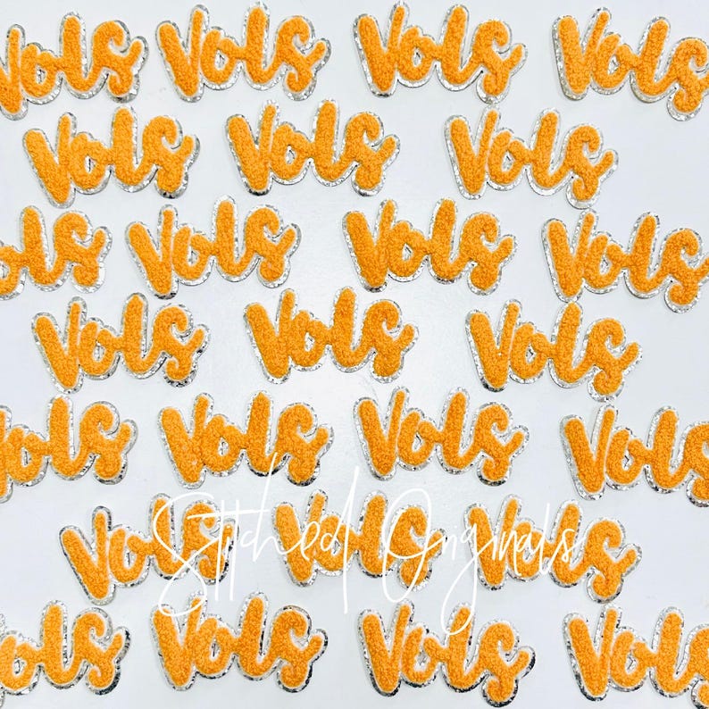May include: A collection of orange and silver iron-on patches with the word "Vols" written in a cursive font.