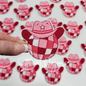 May include: A collection of pink and red plaid iron-on patches featuring a cowboy hat with a star. The patches are in the shape of a rounded square with a slight curve at the top.