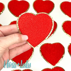 May include: A close-up of a red felt heart with a gold glitter outline. The heart is being held in a hand. The heart is one of many hearts in the image.