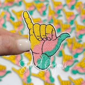 May include: A yellow, pink, and green embroidered iron-on patch of a hand making the shaka sign. The patch is surrounded by other similar patches.
