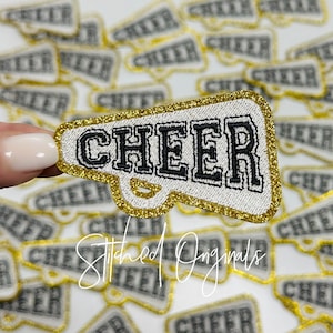 May include: A gold glitter iron-on patch shaped like a megaphone with the word "CHEER" in black outlined letters. The patch is surrounded by other similar patches.