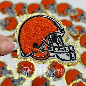 May include: A brown and white embroidered football helmet patch with a gold glitter outline. The patch is made of felt and has a textured surface. The patch is a popular item for fans of the Cleveland Browns.