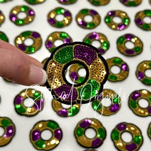 May include: A close-up of a purple, green, and gold sequined donut-shaped patch. The patch is surrounded by other similar patches in various colors.