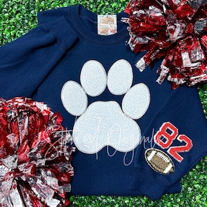 May include: Navy blue sweatshirt with a white sparkly paw print design. The sweatshirt has a red and white football patch on the sleeve with the number 82.