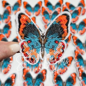 May include: A close-up of a blue, orange, and black butterfly-shaped patch with sequins. The patch is on a white background.