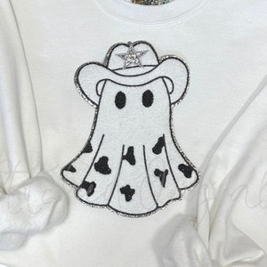 May include: White sweatshirt with a sparkly white ghost wearing a cowboy hat with a silver star on top. The ghost has black cow print on its bottom half.