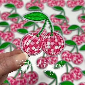 May include: A close-up of a pink and white checkered cherry patch. The cherry is made of felt and has a green stem and leaf. The patch is perfect for adding a touch of whimsy to any project.