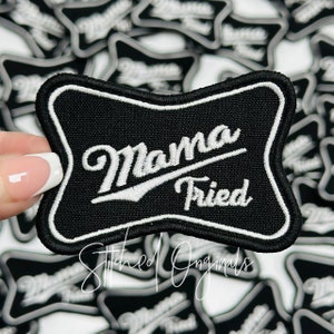 May include: Black and white embroidered patch with the text "Mama Fried" in a white cursive font on a black background. The patch is shaped like a beer bottle label and has a white border.