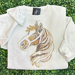 May include: A beige sweatshirt with a sequined horse head design. The horse is brown and white with a gold bridle. The sweatshirt has a crew neck and long sleeves.