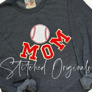 May include: A dark gray sweatshirt with a red, white, and gold baseball-themed "MOM" graphic. The graphic includes a white baseball with red stitching and a gold outline. The text "Stitched Originals" is written in white below the graphic.