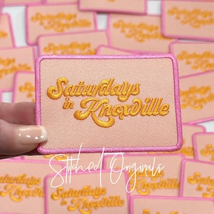 May include: Pink fabric patch with a gold embroidered design that says "Saturdays in Knoxville". The patch has a pink border and is surrounded by other similar patches.