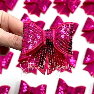May include: A close-up of a hot pink sequined bow with a satin finish. The bow is held in a hand and there are other bows in the background.