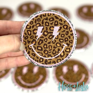 May include: A brown and tan smiley face patch with pink lightning bolts for eyes. The patch is surrounded by a silver glitter trim and has a leopard print pattern.