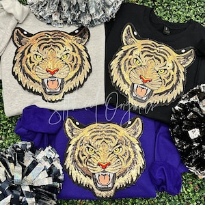 May include: Three sweatshirts in gray, black, and purple, each featuring a large, sequined tiger face. The tiger has gold, black, and brown details with a red nose and open mouth. Pom-poms are visible in the background.