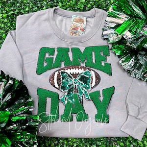 May include: A gray sweatshirt with the words "GAME DAY" in green glitter lettering. A football with a green sequin bow is in the center. The words "Stitched Originals" are written in cursive at the bottom. Green and silver pom-poms are in the background.