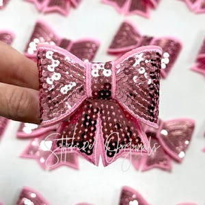 May include: A pink sequined bow with a satin edge. The bow is approximately 2 inches wide and 2 inches tall.