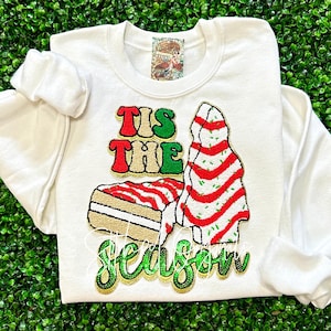 May include: White sweatshirt with a green, red, and gold embroidered design that says "Tis the Season" with a Christmas tree and a slice of cake.