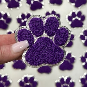 May include: Purple paw print iron-on patch with a silver glitter outline. The patch is made of a soft, fuzzy material.