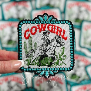 May include: Embroidered patch featuring a cowgirl riding a horse, with the word "COWGIRL" in red. The design includes cacti and a turquoise border. The patch is square with a black border and is approximately 3 inches wide.