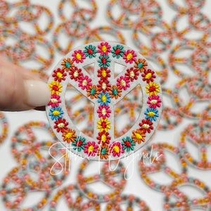 May include: A white peace sign with a colorful flower design embroidered around the edge. The peace sign is made of felt and is approximately 2 inches in diameter.