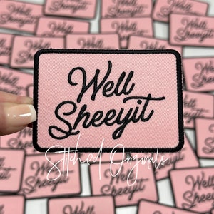 May include: A rectangular pink patch with the words "Well Sheeyit" embroidered in black script. The patch has a black border and is surrounded by other similar patches. The text "Stitched Originals" is also visible.