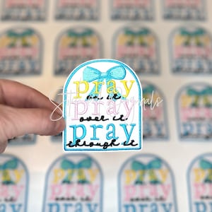 May include: Embroidered patch with the words "Pray over it, pray through it" in yellow, pink, and blue. The patch has a blue bow at the top and a blue border. The patch is held by a hand.