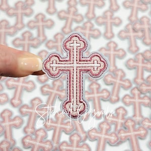 May include: A pink and white embroidered cross with a decorative design. The cross is surrounded by other crosses in the background.