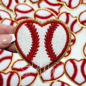 May include: Heart-shaped baseball patch with a white center, red stitching, and a gold glitter border. The patch has a baseball design and is surrounded by other similar patches. The text "Stitched Originals" is visible.